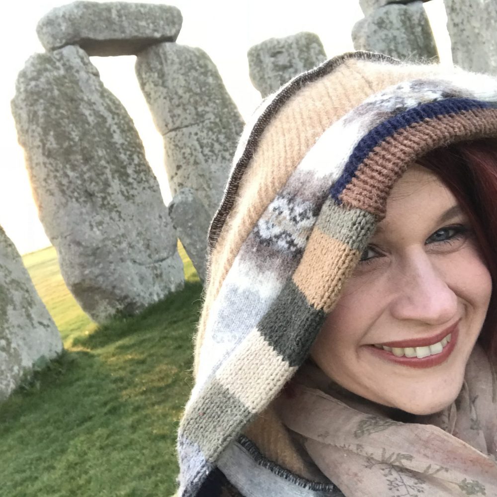 selfie of a smiling woman wearing a hood and with the Stonehenge behind her